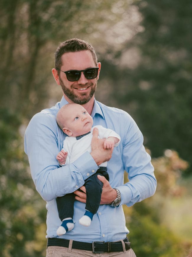 Ein Mann mit Sonnenbrille und hellblauem Hemd lächelt, während er ein Baby in einem weißen Hemd und einer marineblauen Hose im Arm hält. Dieser freudige Moment, der im Freien mit unscharfem Grün aufgenommen wurde, wirkt, als käme er direkt von einer Hochzeit auf Mallorca.