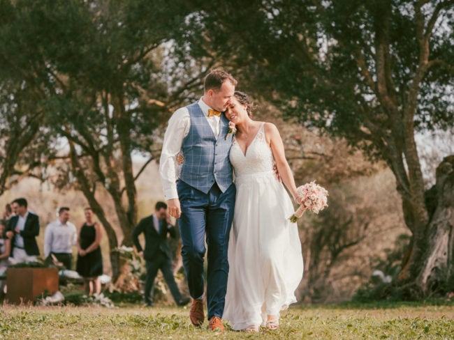 Ein frisch verheiratetes Paar geht nach draußen, lächelt und lehnt sich liebevoll aneinander. Die Braut hält einen Blumenstrauß in der Hand, während die Gäste eine schöne Hochzeit auf Mallorca unter sonnenbeschienenen Bäumen im Hintergrund genießen.
