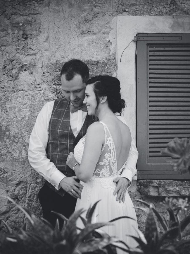 Ein Schwarz-Weiß-Foto von einer Braut und einem Bräutigam, die sich in der Nähe einer Steinmauer und eines Fensters mit Fensterläden umarmen und einander liebevoll anlächeln. Aufgenommen von einem Hochzeitsfotograf Mallorca, trägt die Braut ein Spitzenkleid mit offenem Rücken.