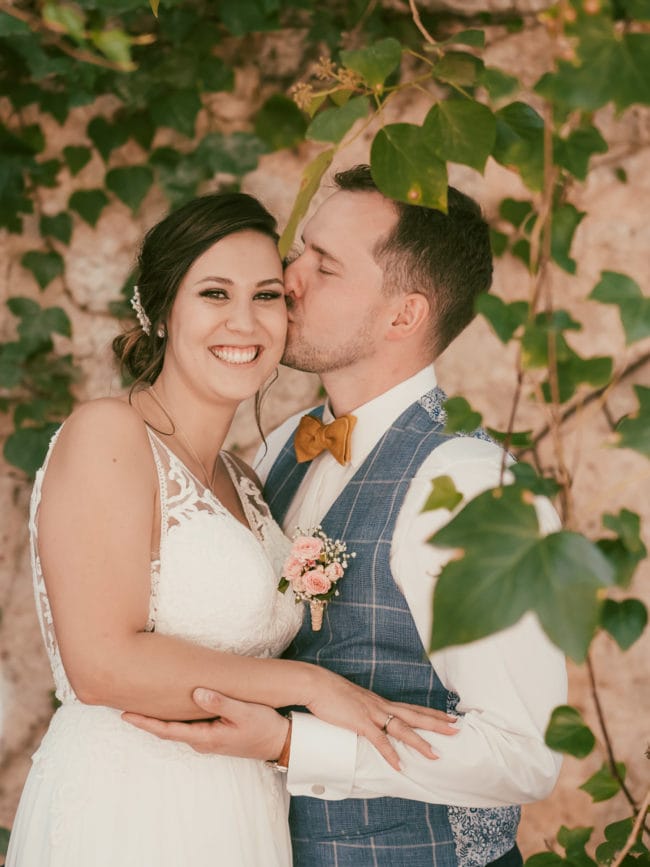 Eine Braut in einem weißen Kleid lächelt, während ihr Bräutigam, der eine blau karierte Weste und eine gelbe Fliege trägt, sie auf die Schläfe küsst. Sie umarmen sich unter blättrigen Weinreben vor einer Steinmauer während ihrer Hochzeit auf Mallorca.