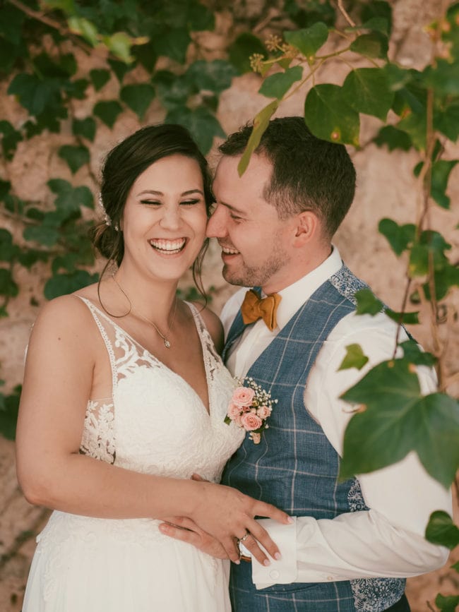 Eine Braut in einem weißen Spitzenkleid und ein Bräutigam in blauer Weste und Fliege lächeln und umarmen sich, während sie an einer begrünten, mit Weinreben bewachsenen Mauer stehen - wunderschön eingefangen von einem Hochzeitsfotografen Mallorca bei ihrer Hochzeit auf Mallorca.