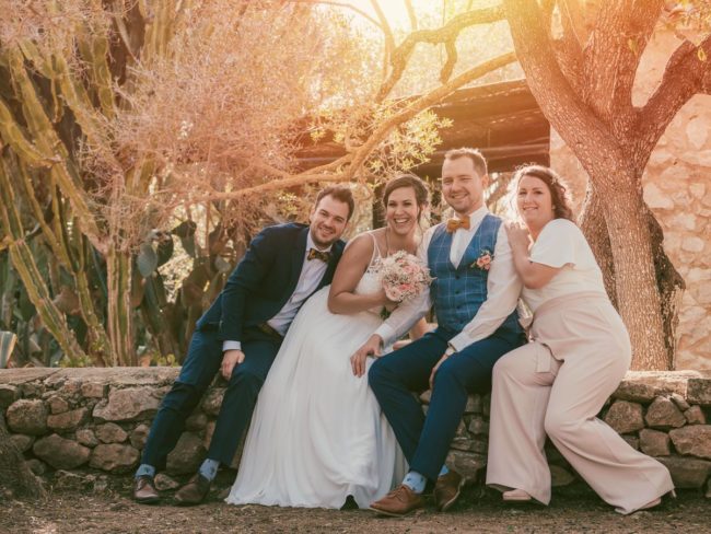 Vier Personen sitzen auf einer Steinmauer im Freien im sanften Sonnenlicht. Zwei Männer in Anzügen und zwei Frauen, eine im Hochzeitskleid mit einem Blumenstrauß in der Hand, lächeln und posieren eng beieinander - und fangen die Freude einer Hochzeit auf Mallorca inmitten üppiger Bäume und Pflanzen ein.
