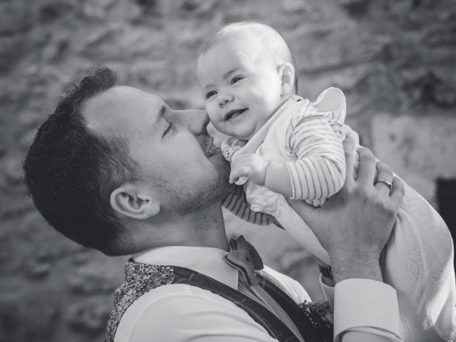 Ein Mann küsst ein lächelndes Baby sanft auf die Wange, während er es in seine Arme nimmt. Beide in formeller Kleidung, teilen sie einen freudigen Moment-perfekt eingefangen von einem Hochzeitsfotograf Mallorca. Das Bild ist in schwarz-weiß.