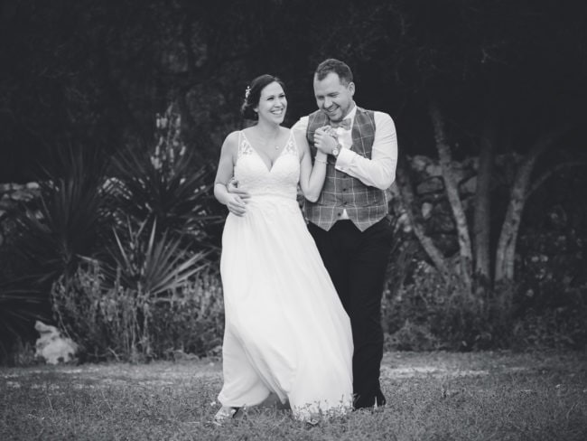 Eine Braut und ein Bräutigam gehen Arm in Arm im Freien, glücklich lächelnd. Aufgenommen von einem Hochzeitsfotograf Mallorca, entfaltet sich ihre Hochzeit auf Mallorca inmitten von Bäumen und Sträuchern, die einen natürlichen, dunklen Hintergrund bilden. Das Bild ist in schwarz-weiß gehalten.