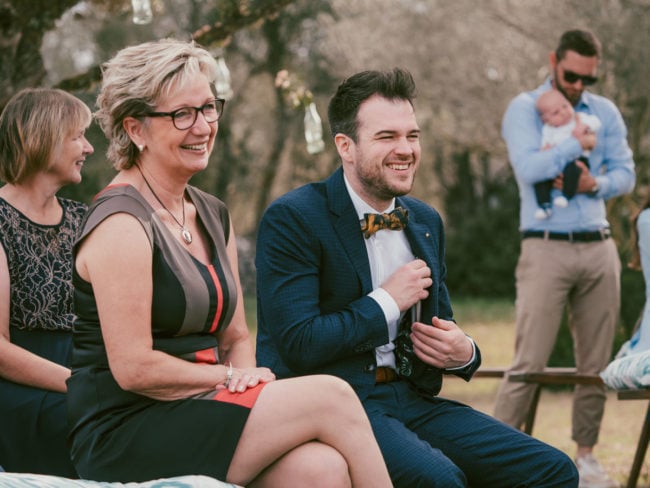 Eine Frau im Kleid und ein Mann im Anzug sitzen lächelnd im Freien auf einer Hochzeit auf Mallorca, eine weitere Frau ist in der Nähe. Im Hintergrund hält ein Mann ein Baby unter einem mit hängenden Glasflaschen geschmückten Baum.