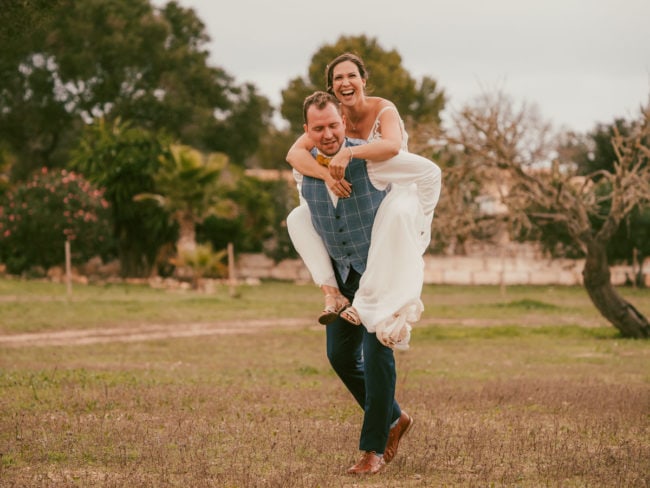 Eine Braut in einem weißen Kleid reitet spielerisch auf dem Rücken ihres Bräutigams, der einen blauen Anzug trägt, auf einer Wiese mit Bäumen im Freien - und fängt damit die Freude einer Hochzeit auf Mallorca ein. Beide lächeln und lachen.