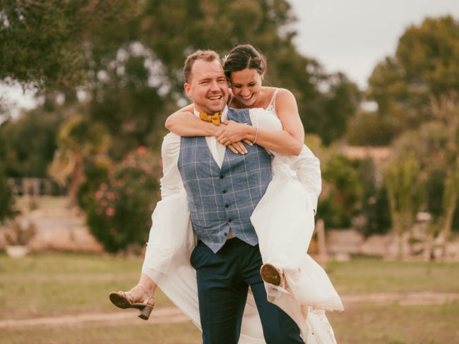 Ein Bräutigam in einer blauen Weste nimmt eine lachende Braut in einem weißen Kleid huckepack, draußen auf einer Wiese mit Bäumen - eingefangen von einem Hochzeitsfotografen Mallorca während einer unvergesslichen Hochzeit auf Mallorca.