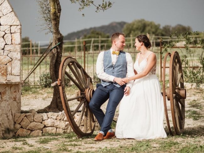 Eine Braut und ein Bräutigam sitzen auf rustikalen Wagenrädern im Freien und lächeln sich während ihrer Hochzeit auf Mallorca an. Der Bräutigam trägt einen blauen Anzug mit gelber Fliege, die Braut ein weißes Kleid, mit Steinen und Grünzeug im Hintergrund.