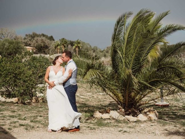 Eine Braut und ein Bräutigam küssen sich im Freien unter einer Palme unter einem schwachen Regenbogen und halten die Freude über ihre Hochzeit auf Mallorca fest. Die helle, natürliche Szene spiegelt die Magie wider, die ein Hochzeitsfotograf Mallorca für immer bewahren kann.