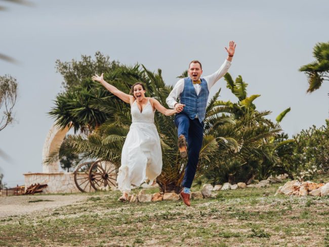 Ein fröhliches Brautpaar, das sich an den Händen hält und lächelt, springt auf seiner Hochzeit auf Mallorca im Freien in die Luft, mit Palmen und einem rustikalen Bogen im Hintergrund.