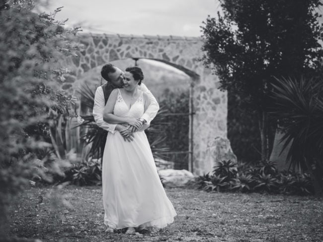 Ein Bräutigam umarmt und küsst seine lächelnde Braut von hinten in einer Gartenlandschaft während ihrer Hochzeit auf Mallorca, beide in Hochzeitskleidung mit Steinbögen und Grün im Hintergrund. Das Schwarz-Weiß-Foto hält diesen besonderen Moment fest.