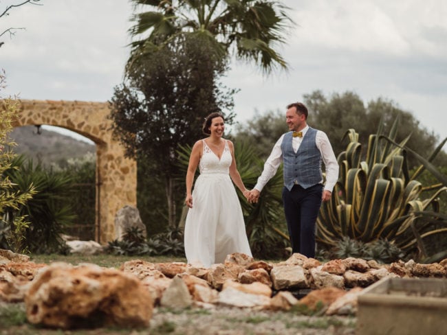 Eine Braut und ein Bräutigam, lächelnd und händchenhaltend, gehen im Freien auf einem felsigen Pfad, umgeben von üppigem Grün und Palmen - perfekt für eine Hochzeit auf Mallorca, wunderschön eingefangen von einem talentierten Hochzeitsfotografen Mallorca.