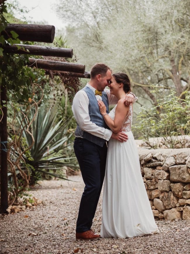 Eine Braut und ein Bräutigam stehen eng beieinander, lächeln und berühren sich an der Stirn, in einem Garten mit Steinmauern und üppigem Grün, der den Zauber einer Hochzeit auf Mallorca einfängt.