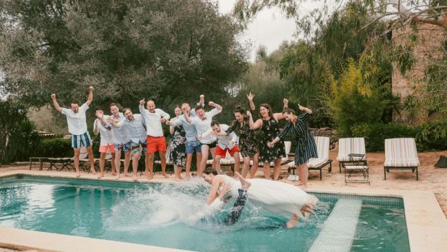 Ein Brautpaar in Hochzeitskleidung stürzt in einen Swimmingpool und spritzt dabei kräftig, während ihre Freunde am Rande des Pools jubeln. So wird die fröhliche Stimmung einer Hochzeit auf Mallorca eingefangen, mit Erinnerungen, die jedem Hochzeitsfotografen Mallorca würdig sind.