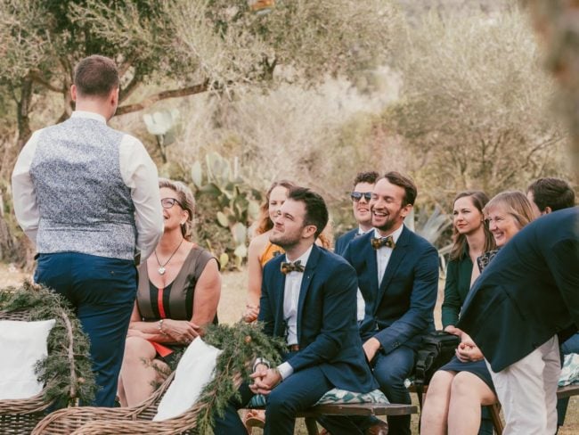 Eine Gruppe gut gekleideter Menschen sitzt im Freien, lächelt und hört einem Mann zu, der anscheinend eine Hochzeit auf Mallorca feiert, mit Bäumen und Grün im Hintergrund - perfekt für jeden Hochzeitsfotografen auf Mallorca.