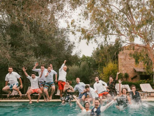 Eine Gruppe von Menschen, die meisten in Badekleidung und einige in Hemden, springt während einer Hochzeit auf Mallorca freudig in einen Swimmingpool, umgeben von Bäumen und Liegestühlen in einer einladenden Umgebung im Freien.
