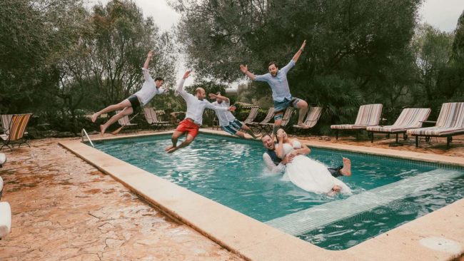 Ein Brautpaar in Hochzeitskleidung fällt während seiner Hochzeit auf Mallorca lachend in einen Pool. Fünf Männer springen hinter ihnen in die Luft. Liegestühle und Bäume umgeben den Pool und tragen zur festlichen Stimmung im Freien bei.