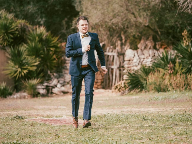Ein Mann im blauen Anzug und mit Fliege geht selbstbewusst auf einer Wiese im Freien, umgeben von Grün und Steinmauern, während das Sonnenlicht durch die Bäume fällt - die perfekte Inspiration für eine Hochzeit auf Mallorca.