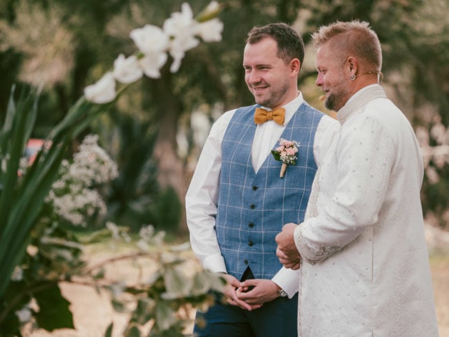 Zwei Männer stehen im Freien auf einer Hochzeit auf Mallorca. Der eine trägt eine blaue Weste mit gelber Fliege und geblümter Boutonniere, der andere, in cremefarbener Stickerei, hält ein Mikrofon. Blumen und Grünzeug umrahmen diesen Moment, der von einem Hochzeitsfotograf Mallorca festgehalten wurde.