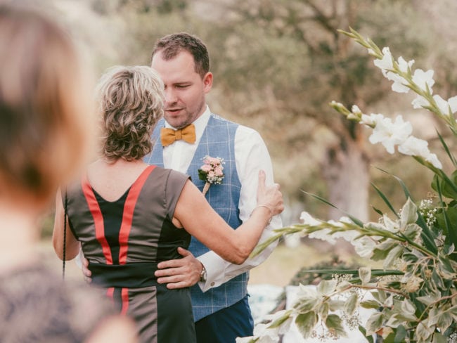 Ein Mann mit blauer Weste und gelber Fliege tanzt mit einer Frau in einem schwarzen Kleid mit roten Streifen auf einer Hochzeit im Freien auf Mallorca, umgeben von Grünzeug und weißen Blumen.