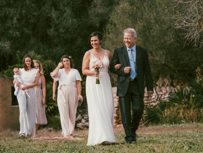 Eine Braut in einem weißen Kleid, die einen Blumenstrauß hält und lächelt, geht Arm in Arm mit einem älteren Mann im Anzug. Vier Frauen, eine mit einem Baby in der Hand, folgen ihnen auf einer Wiese bei einer Hochzeit auf Mallorca.