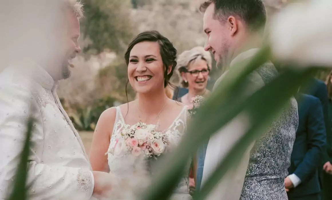 Eine Braut lächelt strahlend, während sie einen Blumenstrauß in der Hand hält und zwischen zwei Männern bei einer Hochzeit im Freien auf Mallorca steht. Andere Gäste sind im verschwommenen Hintergrund zu sehen und Blumen umrahmen den Vordergrund, perfekt eingefangen von einem Hochzeitsfotograf Mallorca.