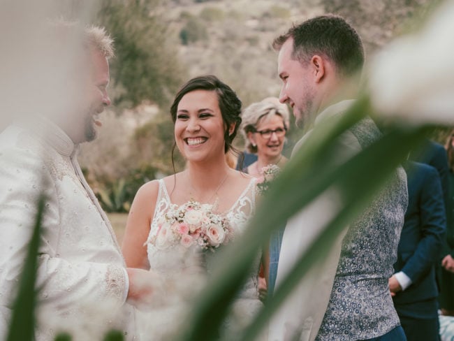 Eine Braut lächelt strahlend, während sie einen Blumenstrauß in der Hand hält und zwischen zwei Männern bei einer Hochzeit im Freien auf Mallorca steht. Andere Gäste sind im verschwommenen Hintergrund zu sehen und Blumen umrahmen den Vordergrund, perfekt eingefangen von einem Hochzeitsfotograf Mallorca.