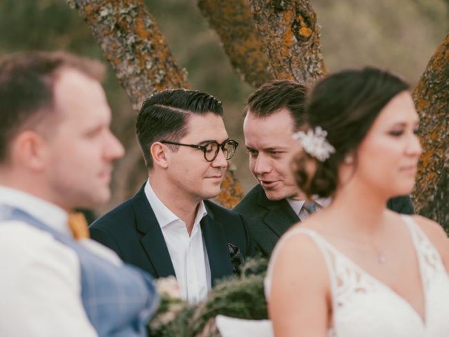 Zwei Männer in Anzügen sitzen im Freien unter Bäumen, einer flüstert dem anderen bei einer Hochzeit auf Mallorca etwas zu. Im Vordergrund sind ein Mann in einer blauen Weste und eine Frau in einem weißen Kleid mit einem Haarschmuck leicht unscharf.