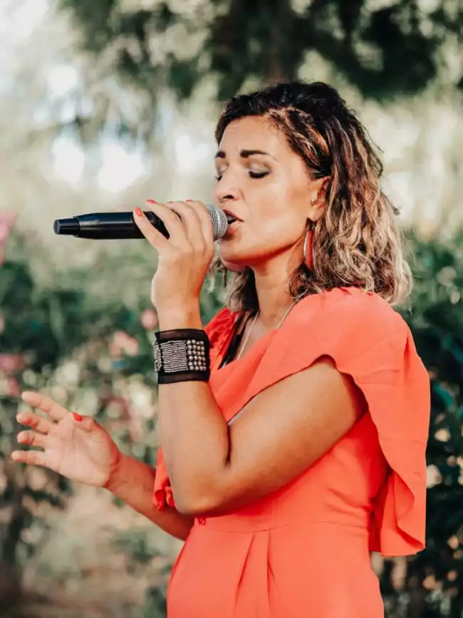 Eine Frau in einem leuchtend orangefarbenen Kleid singt unter freiem Himmel leidenschaftlich in ein Mikrofon, die Augen geschlossen und eine Hand erhoben, umgeben vom Sonnenlicht - und fängt so in jedem Moment den Zauber einer Hochzeit auf Mallorca ein.
