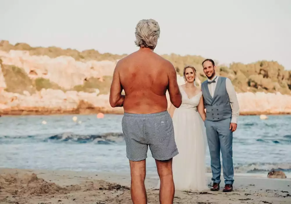 Ein älterer Mann ohne Hemd und in Badehose steht am Strand und blickt auf ein lächelndes Brautpaar, das für ein Foto mit dem Meer und den felsigen Klippen posiert - und so den fröhlichen Geist einer Hochzeit auf Mallorca einfängt.