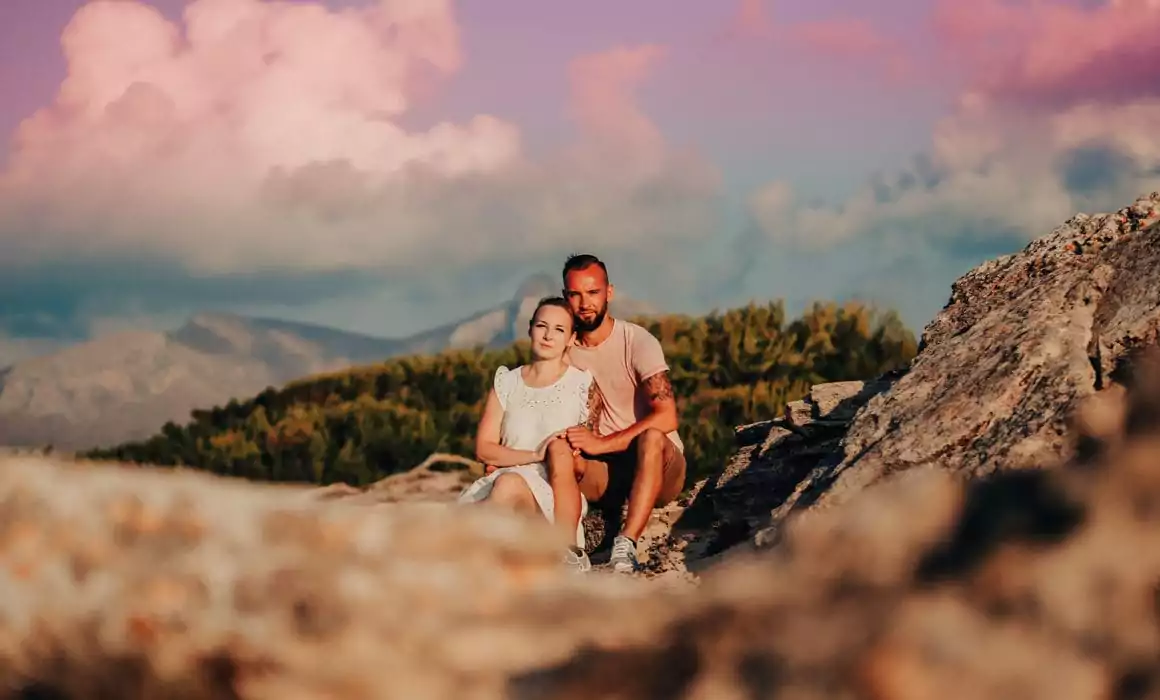 Ein Paar sitzt eng beieinander auf einem felsigen Gelände, während sich ihre Hochzeit auf Mallorca entfaltet, mit üppigen grünen Sträuchern und fernen Bergen hinter ihnen unter einem rosa gefärbten Himmel mit malerischen Wolken.