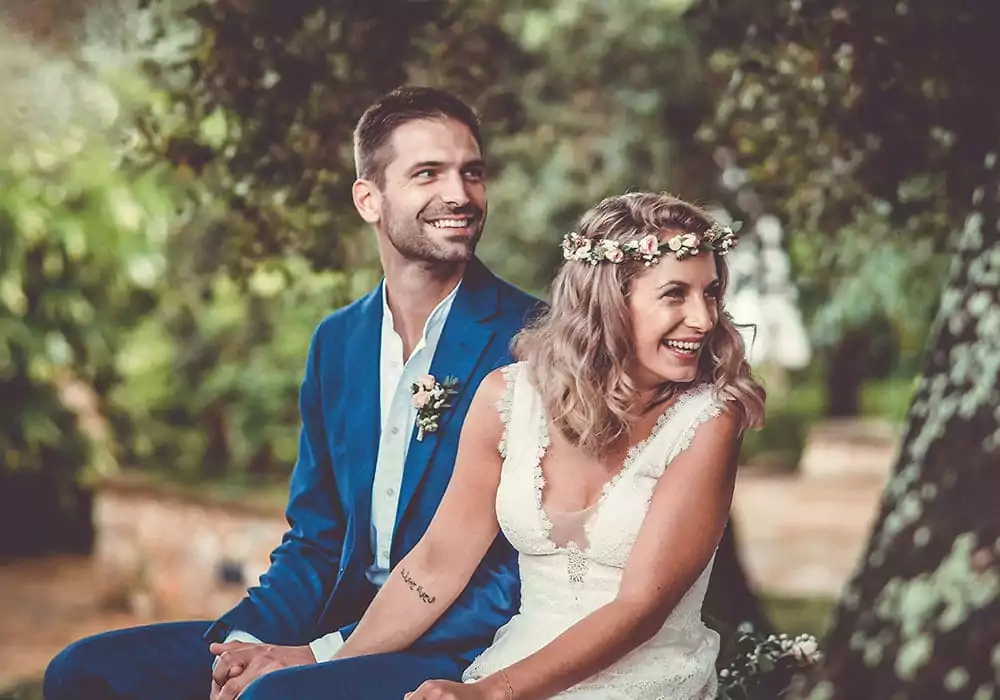 Eine lächelnde Braut in einem weißen Kleid und einer Blumenkrone sitzt neben einem Bräutigam in einem blauen Anzug im Freien, hält Händchen und lacht inmitten von Grün und sanftem Sonnenlicht - perfekte Momente für einen Hochzeitsfotografen Mallorca, um Ihre Hochzeit auf Mallorca festzuhalten.