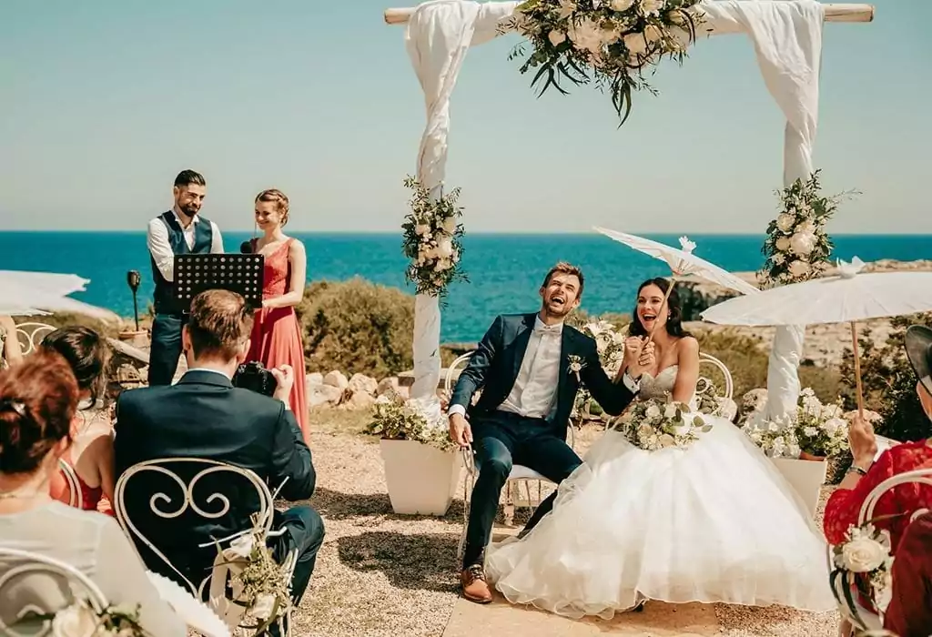 Braut und Bräutigam sitzen lächelnd unter einem Blumenbogen auf ihrer Hochzeit auf Mallorca, mit Gästen auf weißen Stühlen am Meer. Zwei Personen stehen in der Nähe, eine spricht an einem Podium. Das Wetter ist sonnig und hell - perfekt für jeden Hochzeitsfotograf Mallorca.