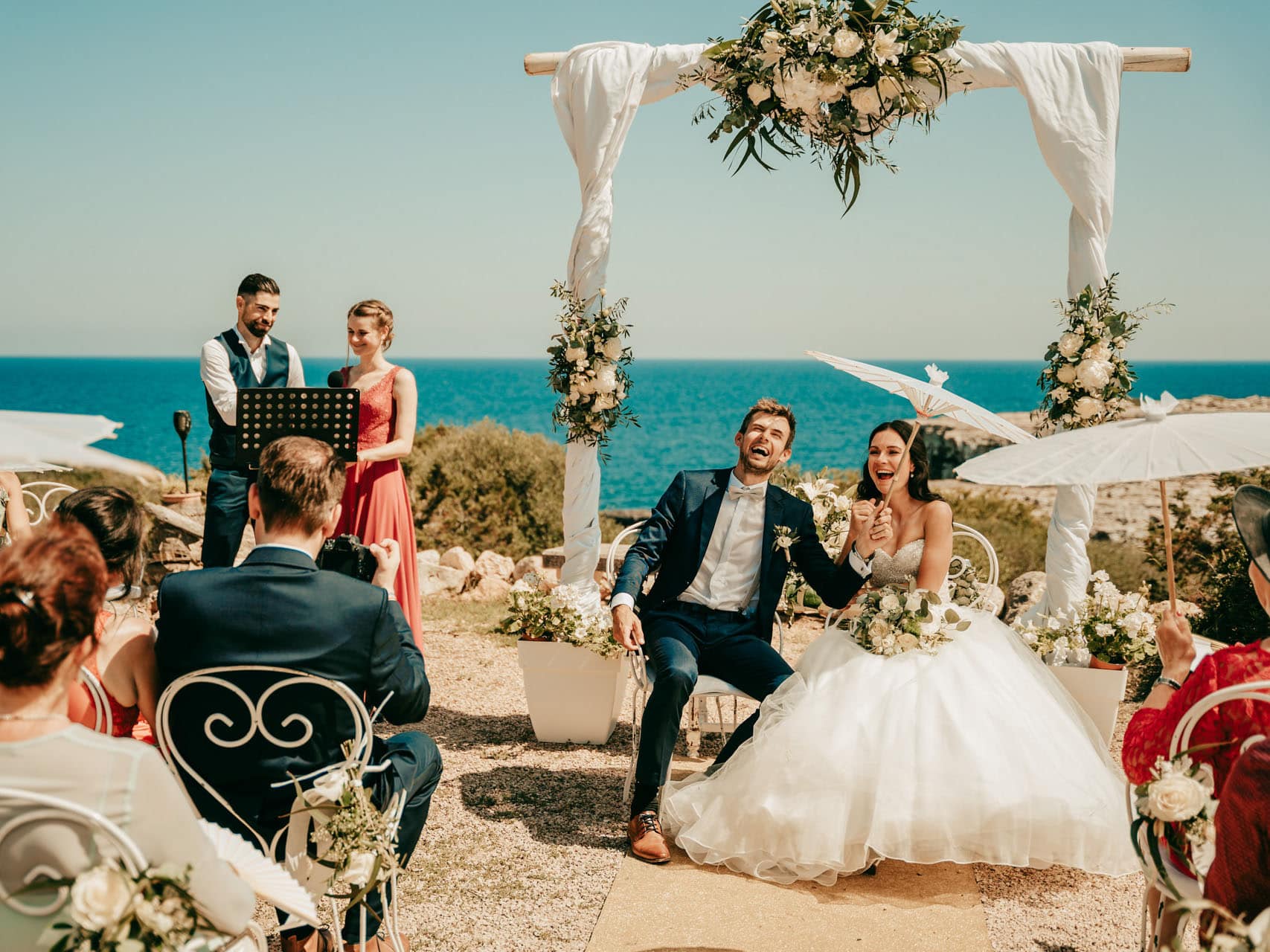 hochzeit am meer auf mallorca Braut und Bräutigam sitzen glücklich unter einem weißen, blumengeschmückten Hochzeitsbogen am Meer, umgeben von Gästen. Die Zeremonie, eine wunderschöne Hochzeit auf Mallorca, findet an einem sonnigen Tag im Freien statt, während der Hochzeitsfotograf Mallorca jeden freudigen Moment festhält.