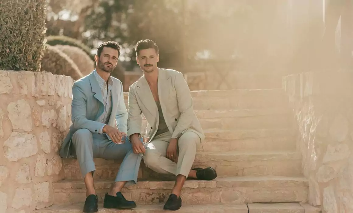 Two men in pastel-colored suits sit closely together on sunlit stone steps, holding hands and looking towards the camera—a moment of love beautifully captured by a Hochzeitsfotograf Mallorca during their Hochzeit auf Mallorca.