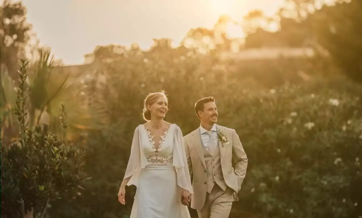 Eine Braut und ein Bräutigam gehen Hand in Hand im Freien bei Sonnenuntergang, lächelnd und zufrieden aussehend. Umgeben von üppigem Grün, ist ihre Hochzeit auf Mallorca wunderschön eingefangen - die Braut in Spitze, der Bräutigam in Beige - von einem talentierten Hochzeitsfotografen Mallorca.