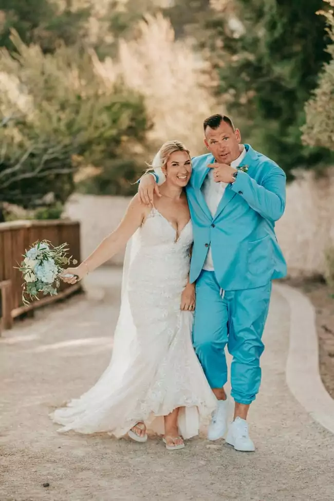 Eine lächelnde Braut in einem weißen Spitzenkleid und Schleier steht Arm in Arm mit einem Bräutigam in einem hellblauen Anzug und weißen Turnschuhen. Ihre verspielte Pose auf einem sonnenbeschienenen Weg, umgeben von Grün, fängt die Freude an einer Hochzeit auf Mallorca ein.