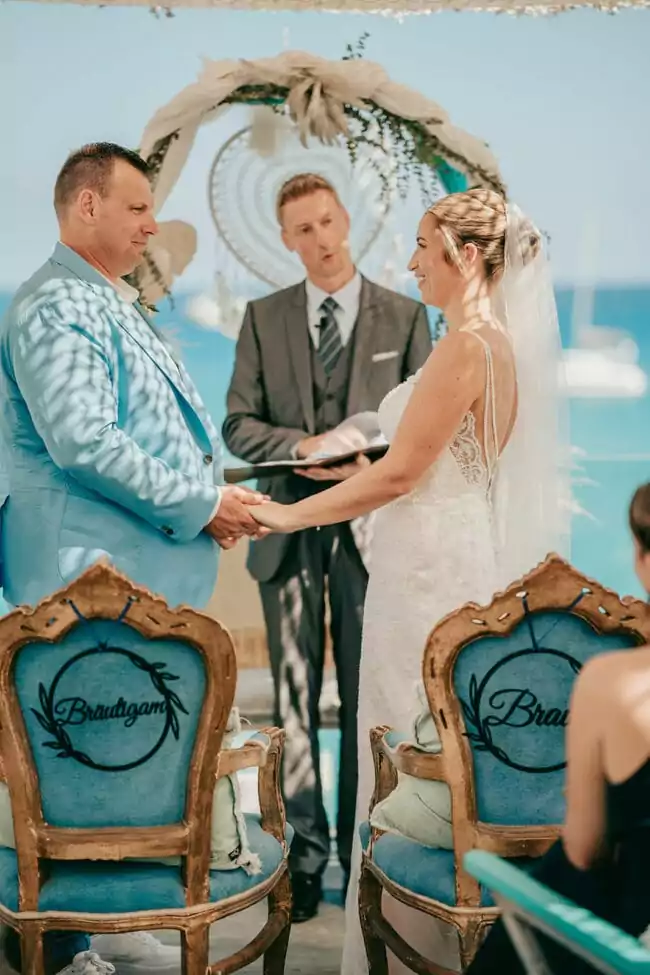 Eine Braut und ein Bräutigam stehen sich während einer Hochzeit im Freien auf Mallorca am Meer händchenhaltend gegenüber, mit einem geschmückten Bogen und einem Geistlichen im Hintergrund. Auf ihren Stühlen stehen 