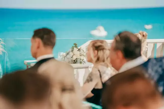 Eine Gruppe von Menschen sitzt vor einem Tisch mit einem weißen Blumenarrangement und blickt auf einen strahlend blauen Ozean mit Booten in der Ferne - eine atemberaubende Hochzeit auf Mallorca, aufgenommen von einem Hochzeitsfotografen Mallorca an einem sonnigen Tag.