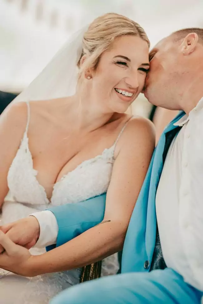 Eine Braut in einem weißen Spitzenkleid lächelt freudig, während ihr Bräutigam in einem hellblauen Anzug sie auf die Wange küsst. Aufgenommen von einem Hochzeitsfotograf Mallorca, halten sie Händchen und strahlen glücklich auf ihrer Hochzeit auf Mallorca.