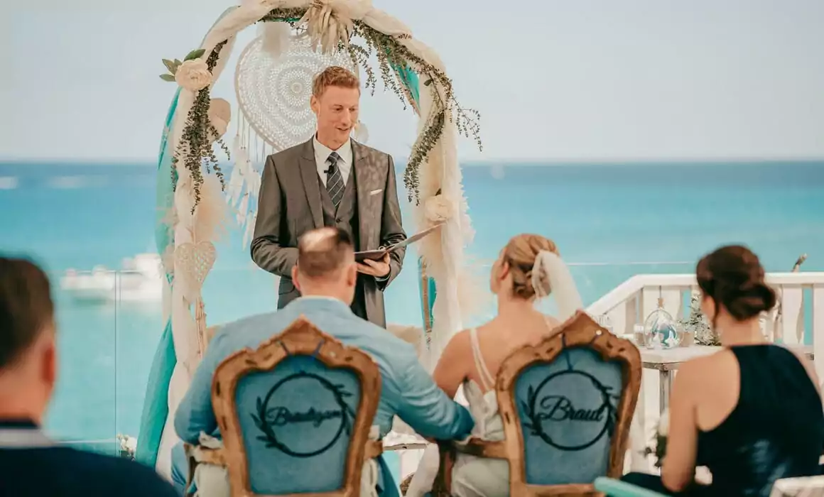 Eine Hochzeitszeremonie am Meer auf Mallorca, mit einem lächelnden Zeremonienmeister unter einem Blumenbogen. Die Braut und der Bräutigam sitzen auf verzierten Stühlen mit der Aufschrift "Bride and Groom", während die Gäste zusehen - perfekt für jede Hochzeit auf Mallorca, die von einem erfahrenen Hochzeitsfotografen Mallorca festgehalten wurde.