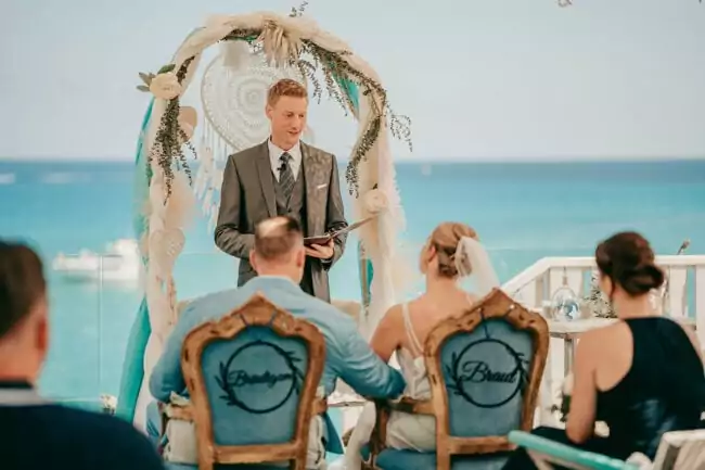 Eine Hochzeitszeremonie am Meer auf Mallorca, mit einem lächelnden Zeremonienmeister unter einem Blumenbogen. Die Braut und der Bräutigam sitzen auf verzierten Stühlen mit der Aufschrift 
