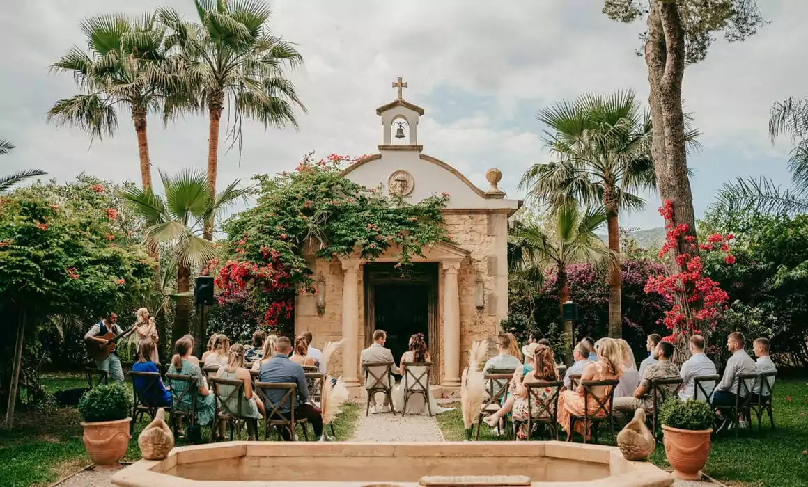 A small outdoor Hochzeit auf Mallorca is taking place in front of a charming stone chapel surrounded by lush greenery, palm trees, and vibrant flowers, as guests are seated facing the chapel under a partly cloudy sky.