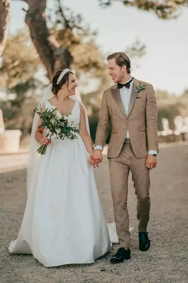 Eine Braut in weißem Kleid und Schleier hält einen Blumenstrauß und geht Hand in Hand mit einem Bräutigam in einem braunen Anzug in der Las Barracas Country Lodge. Sie lächeln sich an und genießen ihre Hochzeit auf Mallorca an einem sonnigen Tag im Freien.