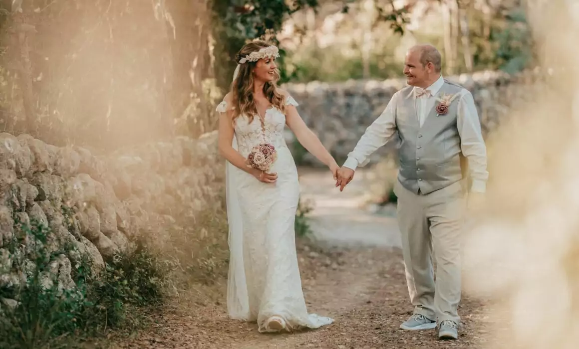 Eine Braut und ein Bräutigam halten sich an den Händen und lächeln sich an, während sie im Freien auf einem sonnenbeschienenen Weg schlendern, der von Steinmauern und Grün umgeben ist - perfekt, um den Zauber einer Hochzeit auf Mallorca einzufangen, wobei jedes Detail von einem Hochzeitsfotografen Mallorca kunstvoll festgehalten wird.