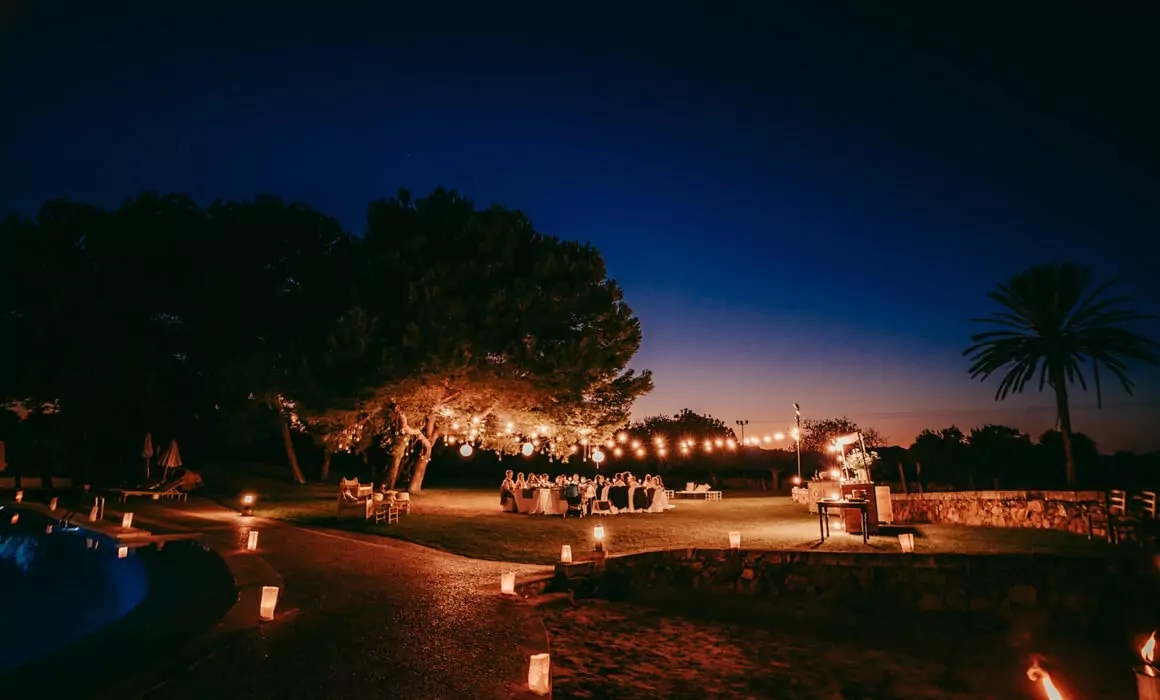An outdoor evening Hochzeit auf Mallorca with string lights hanging from trees, guests at tables, and lanterns lining walkways. The warmly lit scene unfolds beneath a dark blue sky with silhouettes of trees and a palm tree in the background.
