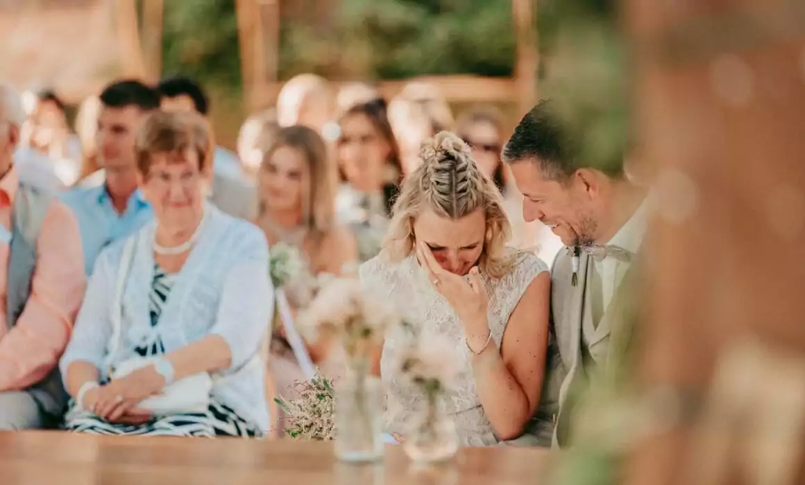 A bride wipes away tears while sitting next to the groom during a Hochzeit auf Mallorca, surrounded by smiling guests in soft, natural light.