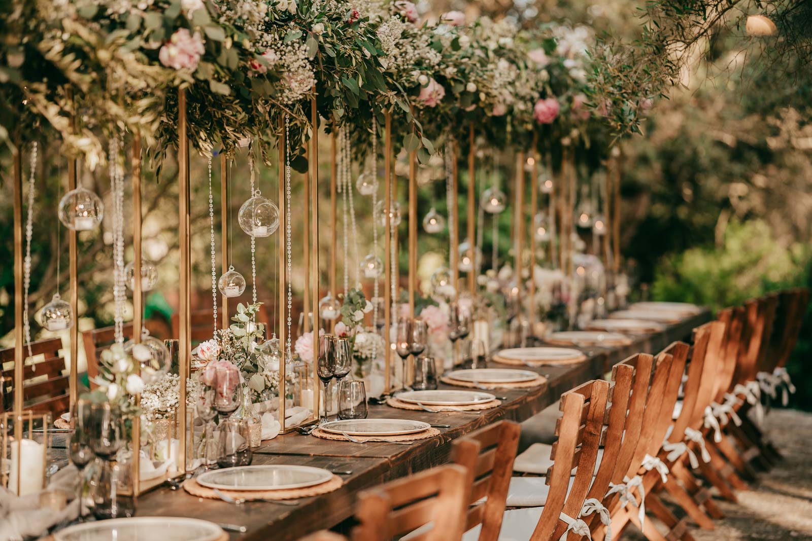 Ein langer, rustikaler Tisch im Freien ist für eine Veranstaltung mit Holzstühlen, Glastellern und eleganten Blumenkränzen gedeckt. Hängende Glaskugeln und üppiges Grün verleihen der natürlichen Gartenumgebung einen romantischen Touch.