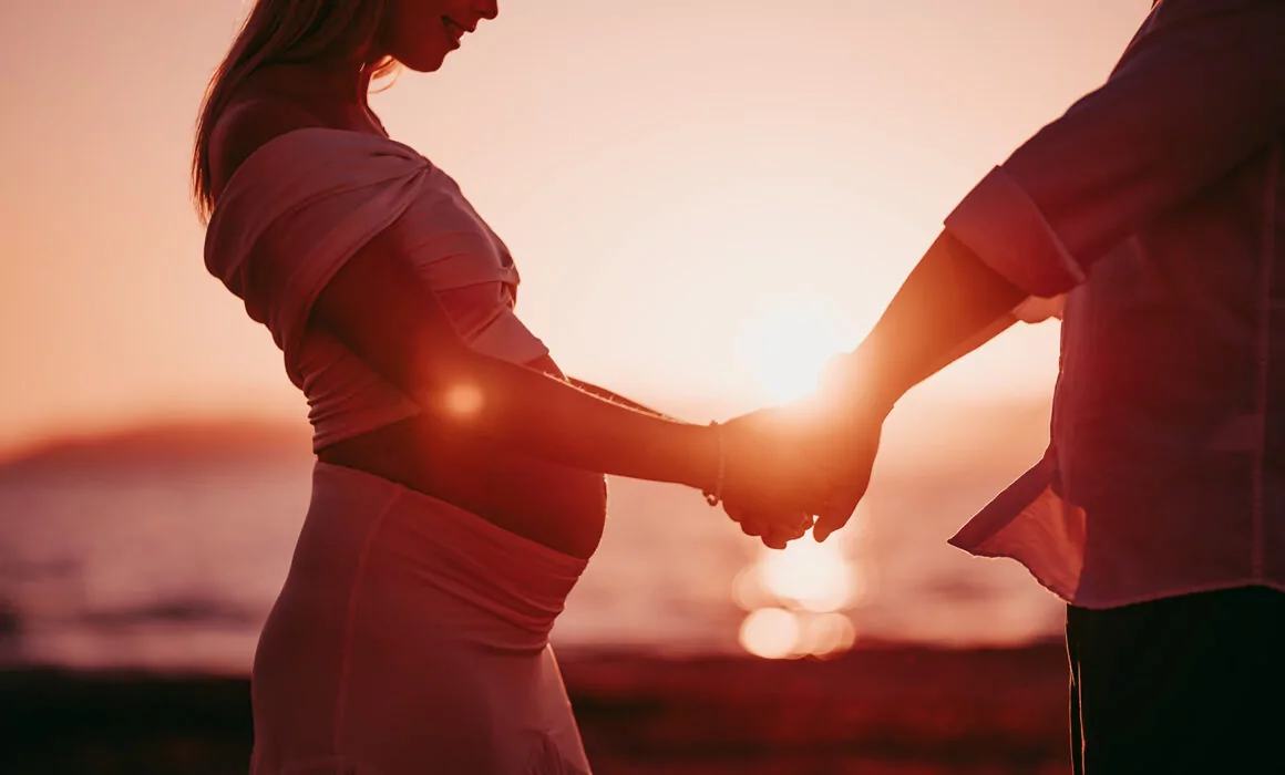 Eine schwangere Frau in einem weißen Kleid hält mit ihrem Partner am Strand während des Sonnenuntergangs Händchen, das warme Licht zeichnet die Silhouetten der beiden - eingefangen von einem Hochzeitsfotografen Mallorca für eine unvergessliche Hochzeit auf Mallorca.