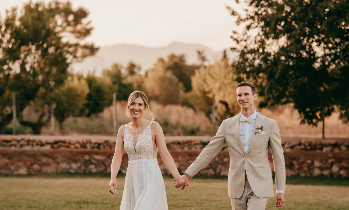 Eine Braut und ein Bräutigam, gekleidet in ein weißes Hochzeitskleid und einen hellbeigen Anzug, halten sich an den Händen und lächeln, während sie draußen auf einem grasbewachsenen Rasen mit Bäumen und Bergen im Hintergrund spazieren gehen - die perfekte Inspiration für eine Hochzeit auf Mallorca.